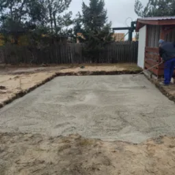 Wyrównywanie świeżo wylanego betonu pod fundamenty z użyciem grabi na działce z drewnianym ogrodzeniem i budynkiem gospodarczym w tle.