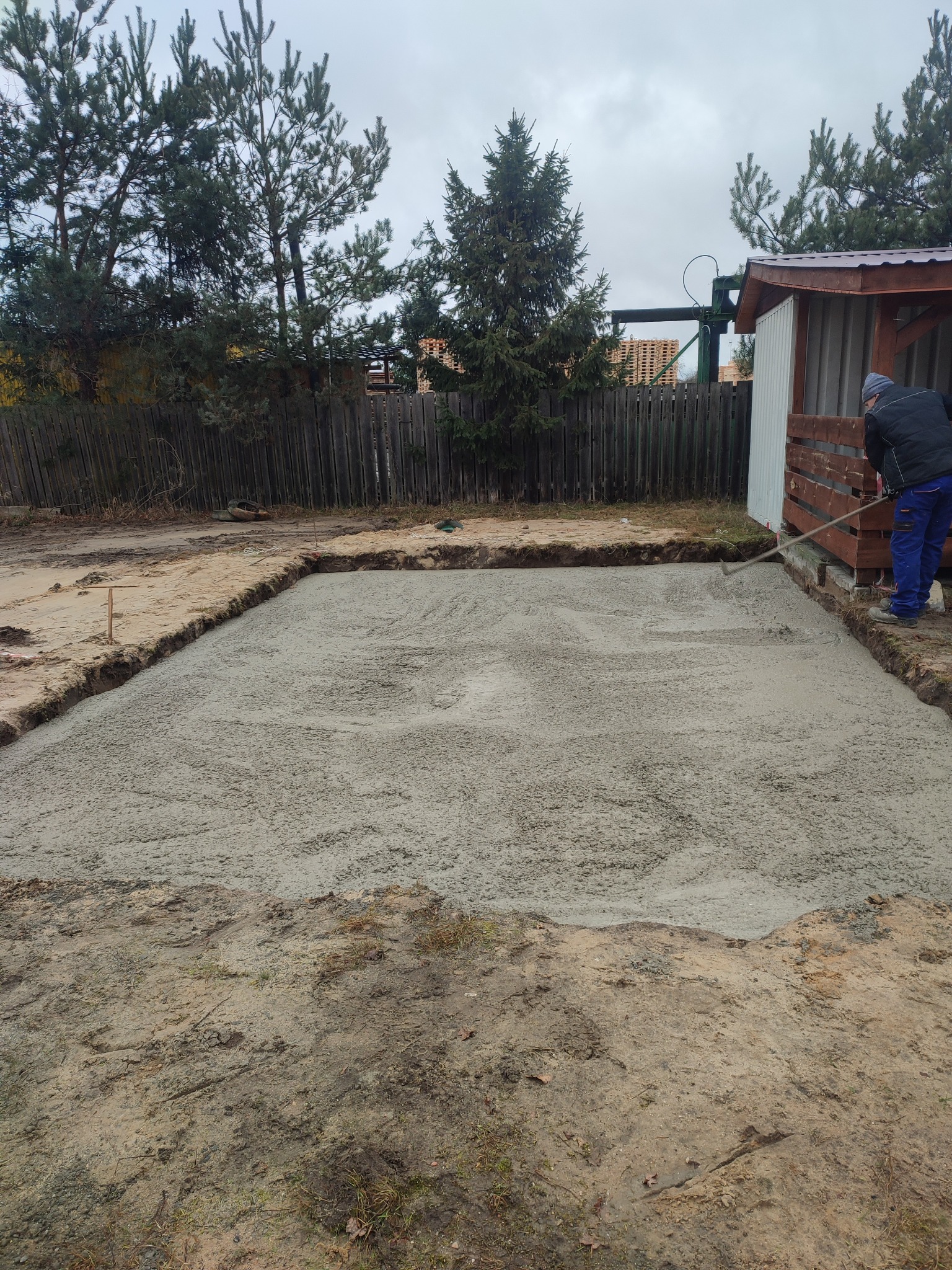 Wyrównywanie świeżo wylanego betonu pod fundamenty z użyciem grabi na działce z drewnianym ogrodzeniem i budynkiem gospodarczym w tle.