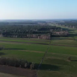 Szeroki kadr z drona na pola uprawne, lasy i łąki, z widocznymi śladami po przejazdach maszyn rolniczych i małymi zbiornikami wodnymi.