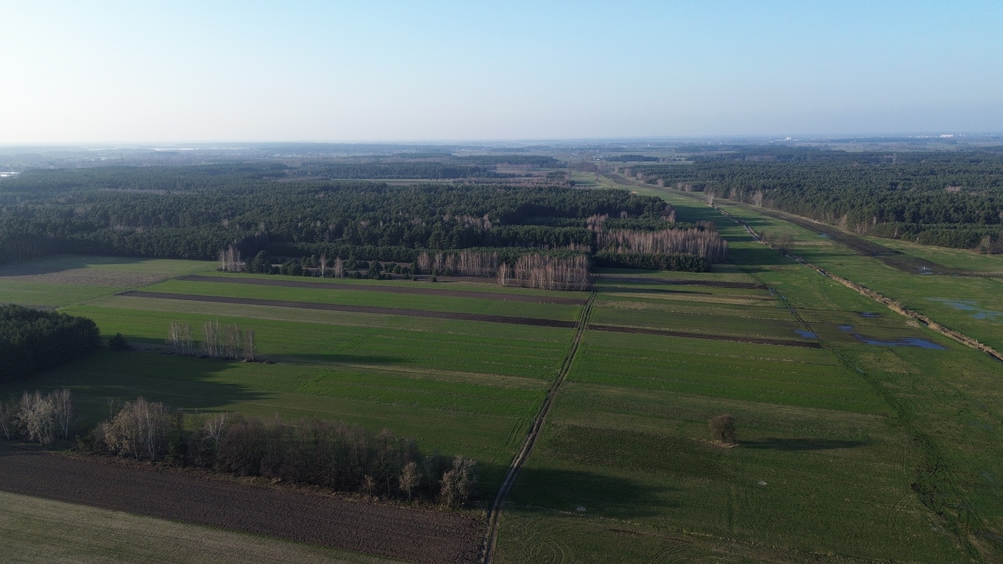 Szeroki kadr z drona na pola uprawne, lasy i łąki, z widocznymi śladami po przejazdach maszyn rolniczych i małymi zbiornikami wodnymi.