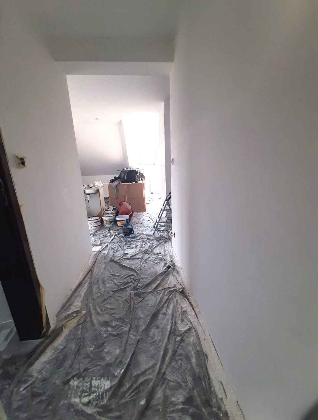 Przygotowanie do malowania: podłoga zabezpieczona folią, widoczne puszki z farbą i drabina w tle, świeżo pomalowane ściany na biało.