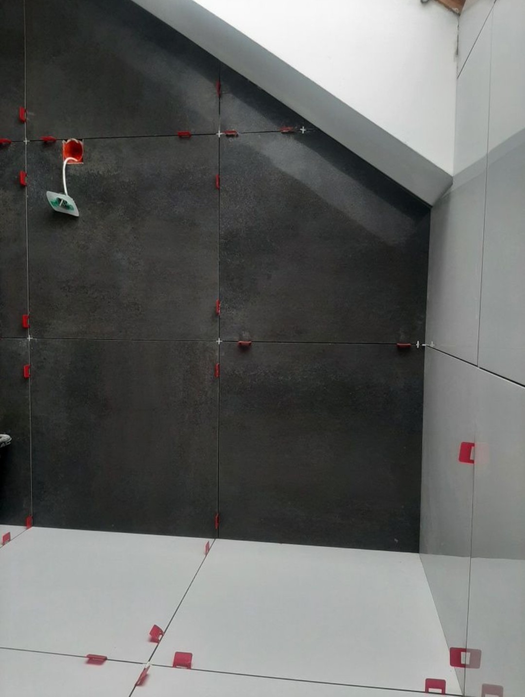 Układanie ciemnoszarych, wielkoformatowych płytek na ścianie ze skosem, z użyciem czerwonych klipsów poziomujących; widoczna puszka elektryczna z wystającym przewodem.