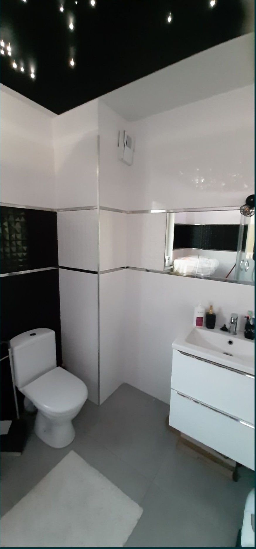 Nowoczesna łazienka z czarno-białymi płytkami, podwieszaną toaletą i sufitem imitującym rozgwieżdżone niebo.