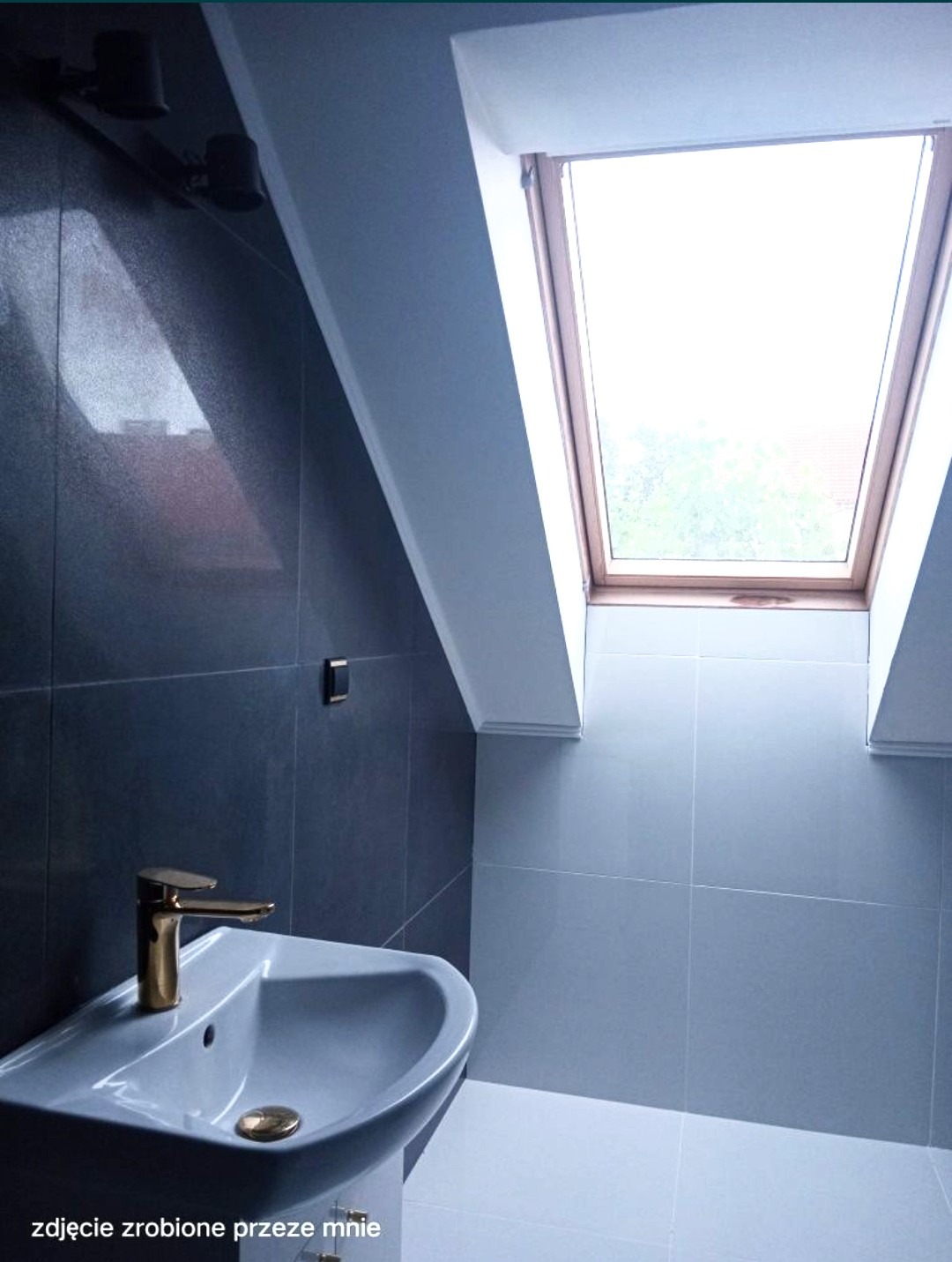 Szara łazienka na poddaszu z umywalką nablatową i złotą baterią, oświetlona oknem dachowym.