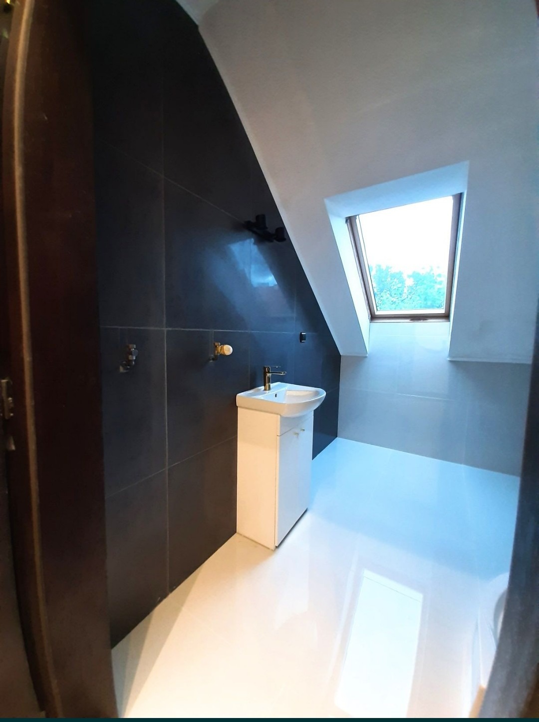 Wykończona łazienka na poddaszu z czarnymi płytkami ściennymi, białą podłogą z połyskiem, umywalką z szafką i oknem dachowym.