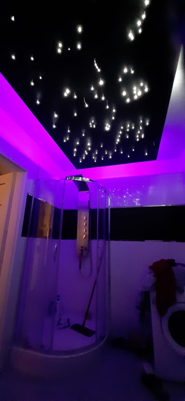 Nowoczesna łazienka z sufitem napinanym imitującym rozgwieżdżone niebo, oświetlonym fioletowym światłem, z kabiną prysznicową i pralką w tle.