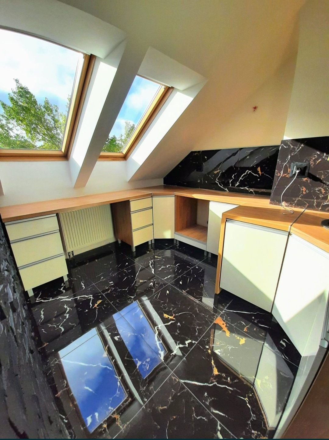 Nowoczesne wnętrze poddasza z czarną marmurową podłogą odbijającą światło z okien dachowych, zabudowa meblowa w jasnych odcieniach z drewnianymi blatami, ściana wykończona płytami z czarnego marmuru.