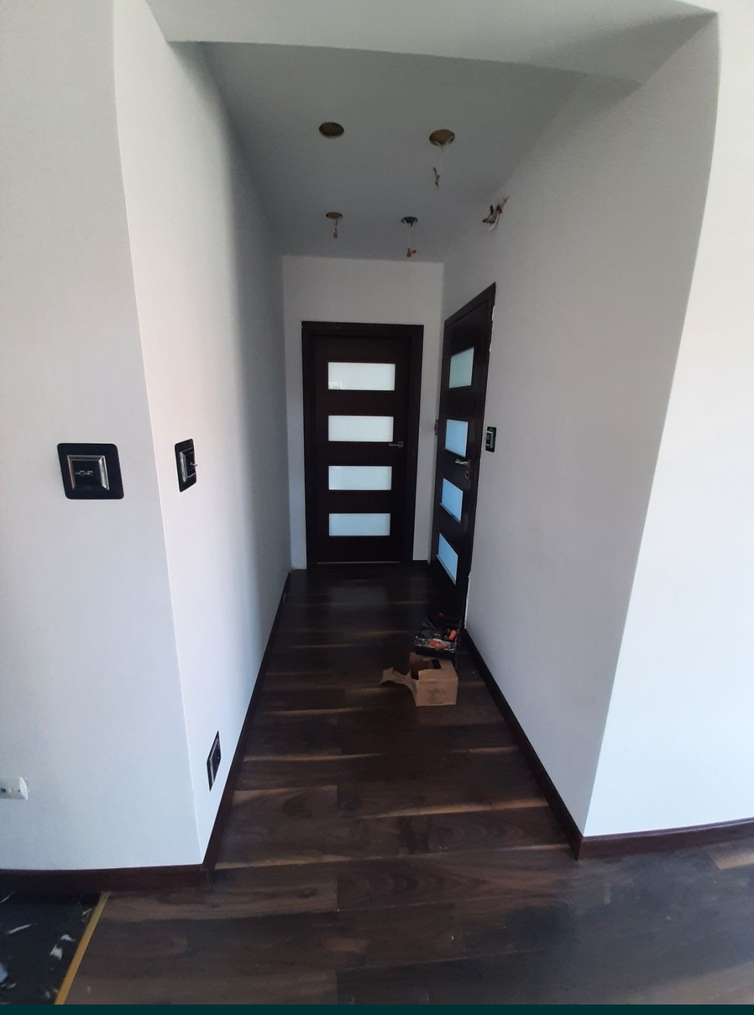 Korytarz z ciemną drewnianą podłogą, białymi ścianami i dwiema ciemnymi drzwiami z przeszkleniami; widoczne narzędzia i karton na podłodze, niedokończone oświetlenie sufitowe.