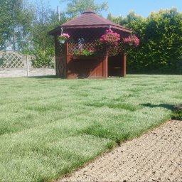 ZieleńPlus - Mężczyzna w kapeluszu układa rolki trawy na przygotowanym gruncie, w tle altana z kwiatami, ogrodzenie i żywopłot.