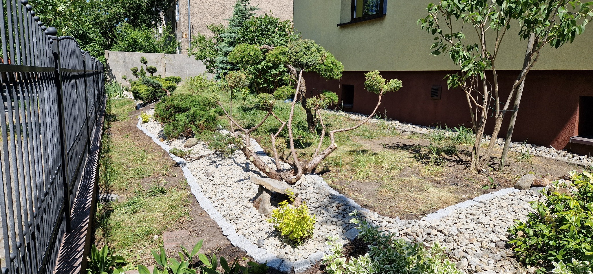 Ogród po niwelacji terenu z kamiennymi ścieżkami, ozdobnymi drzewami i ogrodzeniem z kutego żelaza w słoneczny dzień.
