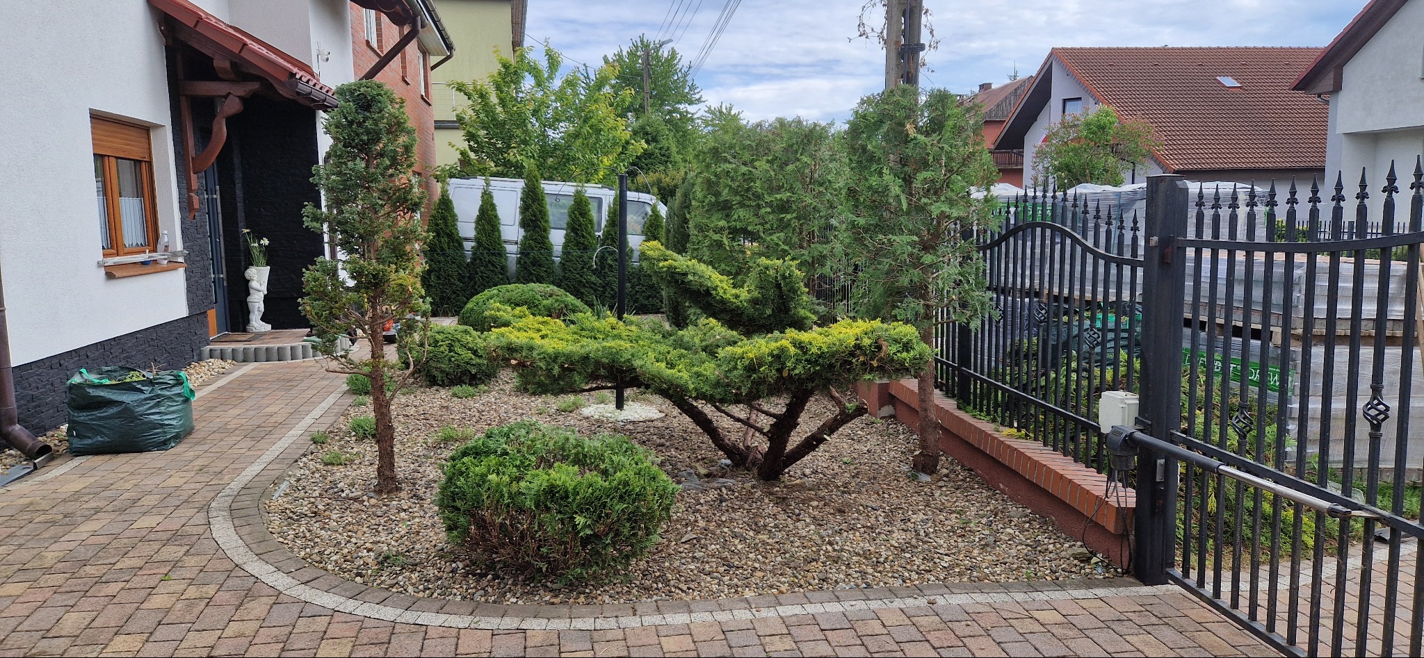 Ogród przydomowy z ozdobnymi krzewami i drzewami iglastymi, wysypany drobnym kamieniem, z widocznym workiem z odpadami ogrodowymi i fragmentem automatycznej bramy w tle.