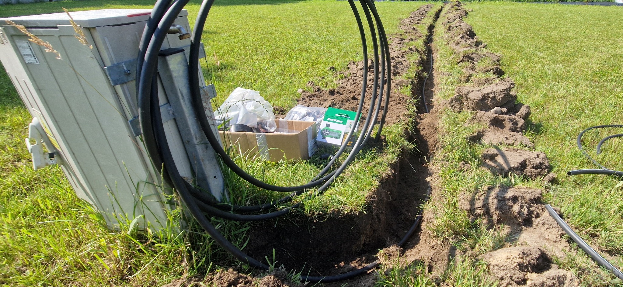 Wykopany rów w trawie z ułożonymi przewodami, prawdopodobnie do systemu nawadniania. Widoczna skrzynka sterująca i akcesoria montażowe.