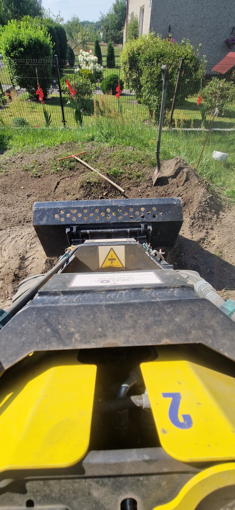 Widok z perspektywy operatora na zagęszczarkę gruntu podczas prac ziemnych w ogrodzie. W tle widoczny ogród z kwiatami i budynek.