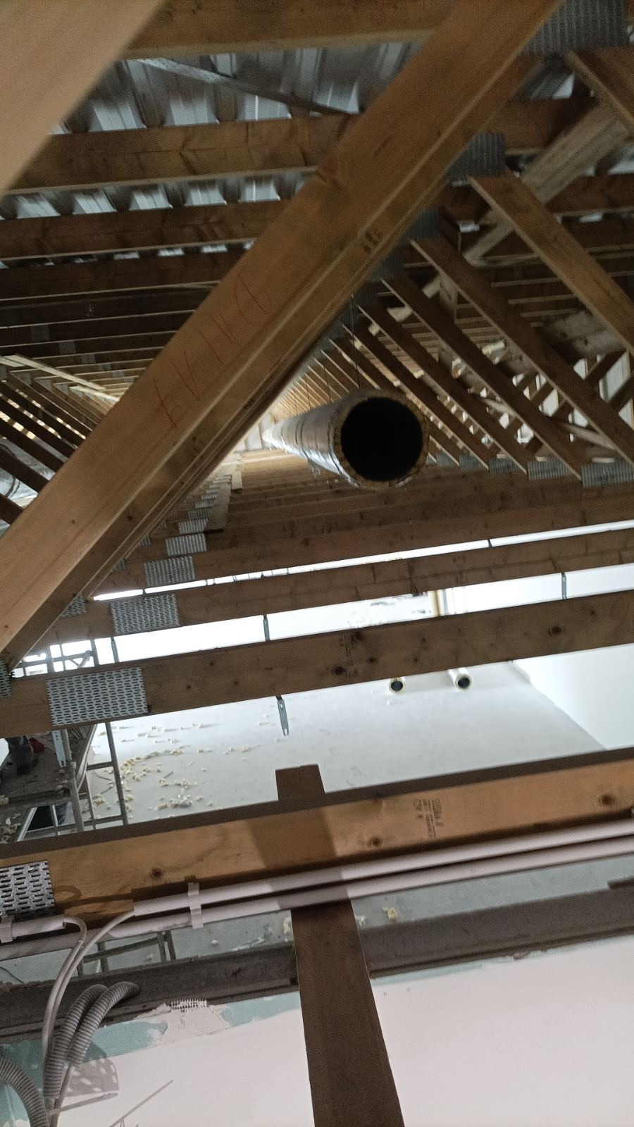 Wnętrze poddasza w trakcie prac wykończeniowych, widoczny fragment systemu wentylacyjnego z okrągłym kanałem poprowadzonym między drewnianymi elementami konstrukcji dachu.
