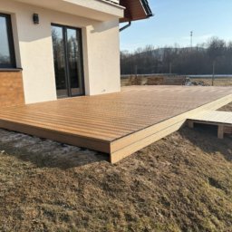 Upwood & lifestyle - Nowoczesny taras kompozytowy z niskimi schodami z boku, przylegający do jasnego budynku z ciemnymi oknami, widok na łąkę i las w tle.