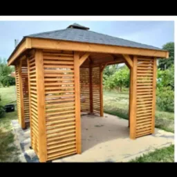 Drewniana konstrukcja z poziomymi listwami, prawdopodobnie altana lub pergola, z ciemnym dachem, na betonowej podstawie, otoczona zielenią.