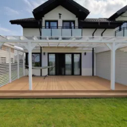 Taras z deski kompozytowej, zadaszony białą konstrukcją drewnianą i przezroczystym poliwęglanem. Widok na dom z balkonami i zielony trawnik.