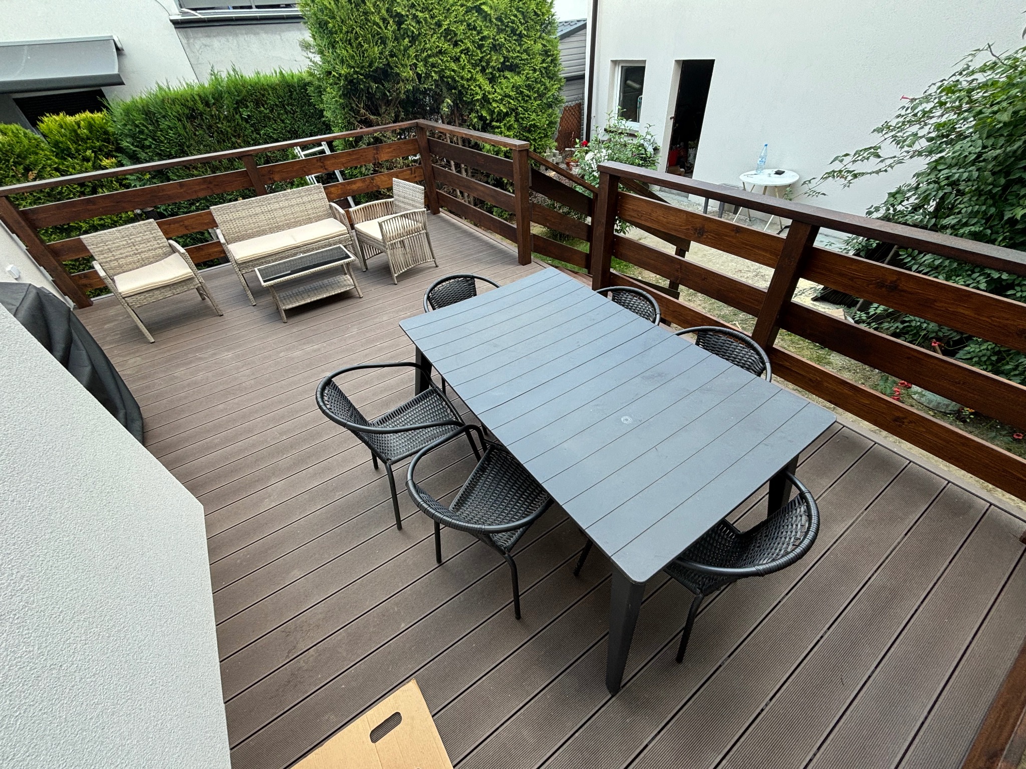 Zewnętrzny taras z szarej deski kompozytowej, wyposażony w stół z krzesłami z plecionki oraz rattanowy zestaw wypoczynkowy, otoczony drewnianą balustradą.