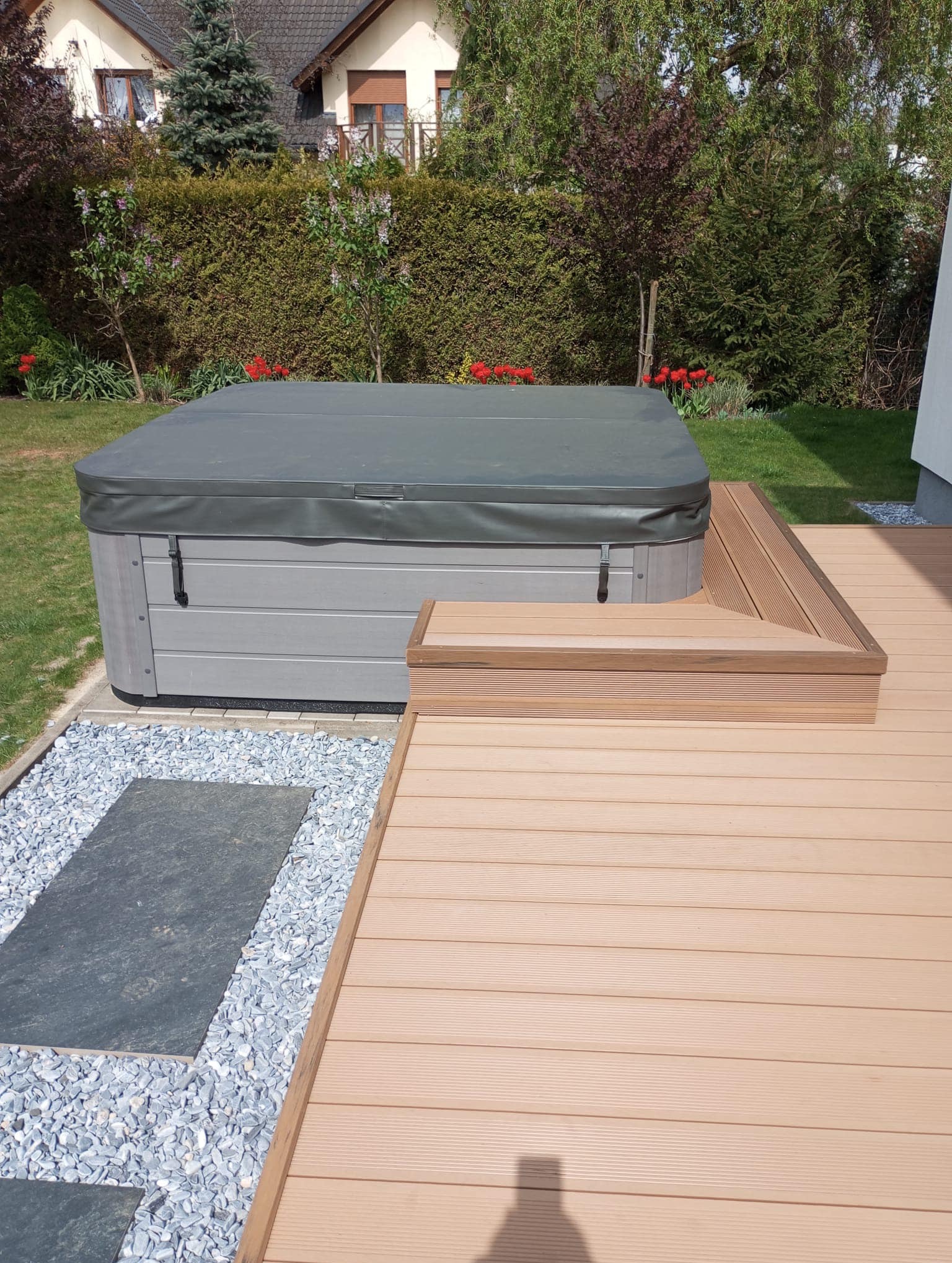 Zadaszone jacuzzi obudowane jasną deską, z niskimi schodkami i ścieżką z kamieni na trawniku w ogrodzie.