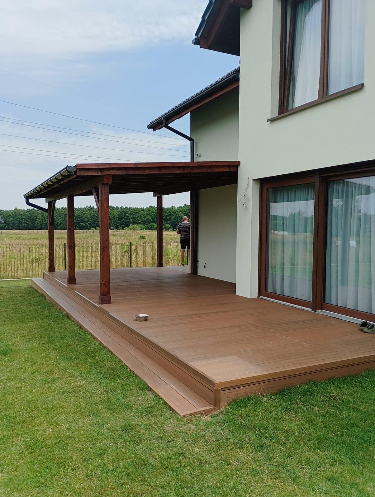 Brązowy taras z kompozytu z zadaszeniem na drewnianych słupach przy domu z dużymi oknami przesuwnymi, widok na łąkę w tle.