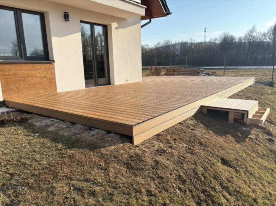 Nowoczesny taras kompozytowy z niskimi schodami z boku, przylegający do jasnego budynku z ciemnymi oknami, widok na łąkę i las w tle.