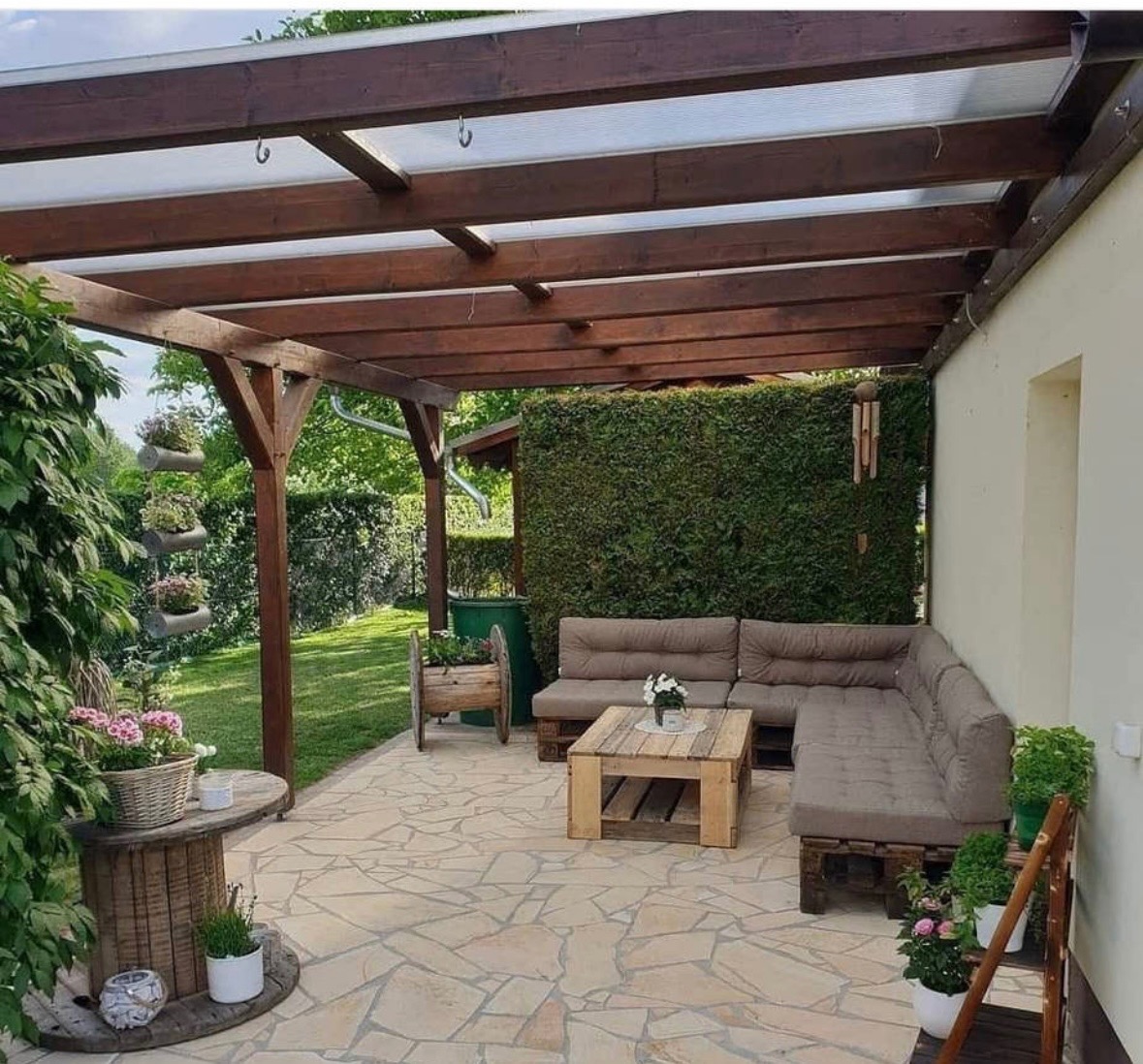 Zadaszona konstrukcja z drewna na tarasie z meblami ogrodowymi z palet, dekoracjami roślinnymi i wiatraczkiem.