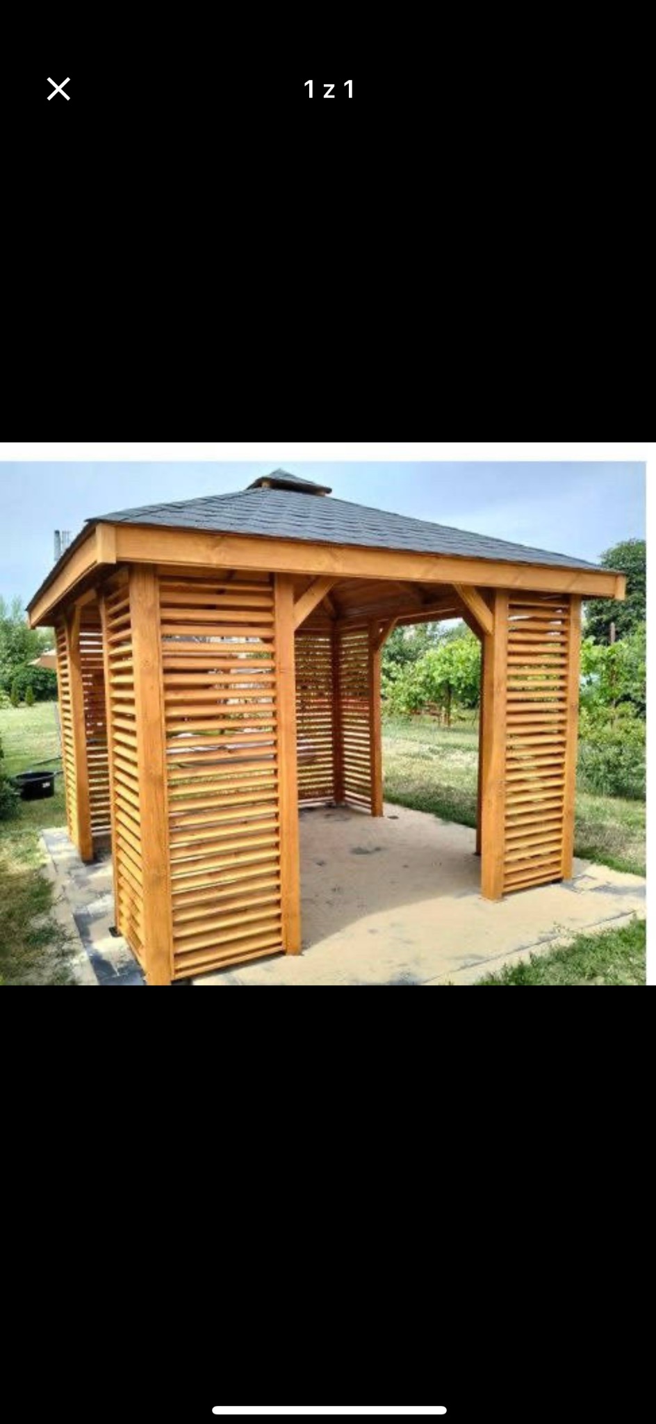 Drewniana konstrukcja z poziomymi listwami, prawdopodobnie altana lub pergola, z ciemnym dachem, na betonowej podstawie, otoczona zielenią.