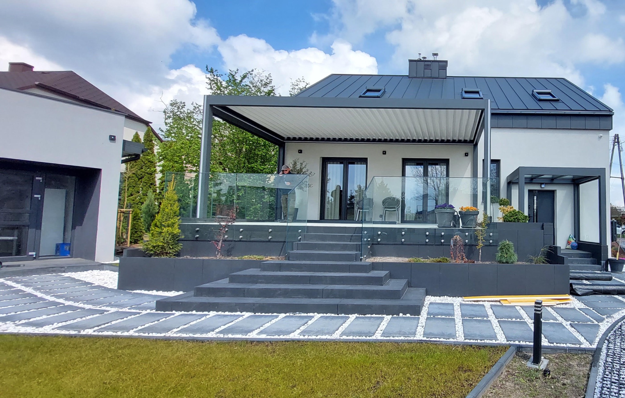 Nowoczesna, antracytowa pergola z lamelami na tarasie. Szklana balustrada, podwyższony taras z schodami. Otoczenie domu z elementami dekoracyjnymi i zielenią.