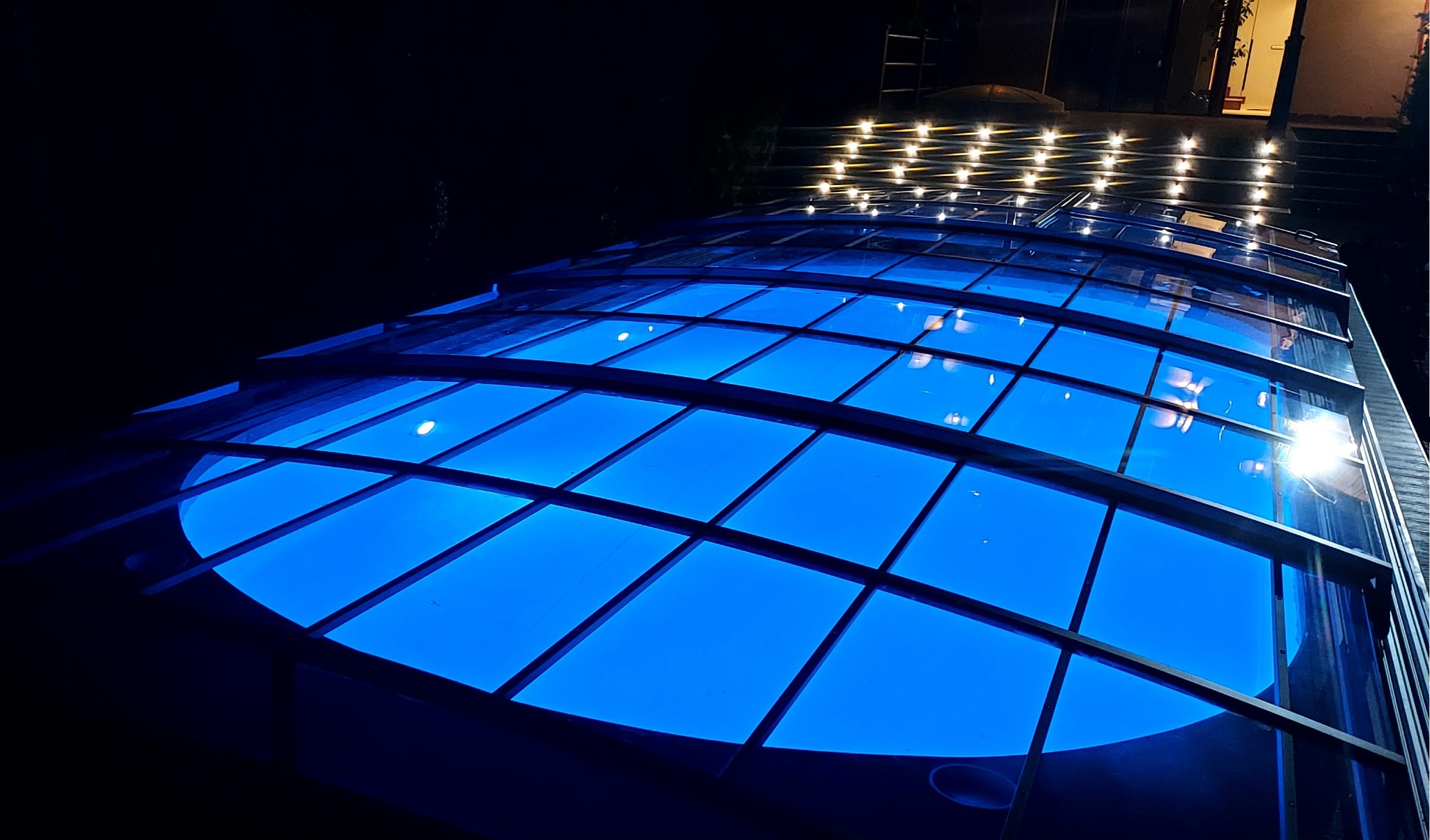 Nocne ujęcie z dołu na zadaszenie basenu z niebieskim oświetleniem, widoczne lampki dekoracyjne w tle.