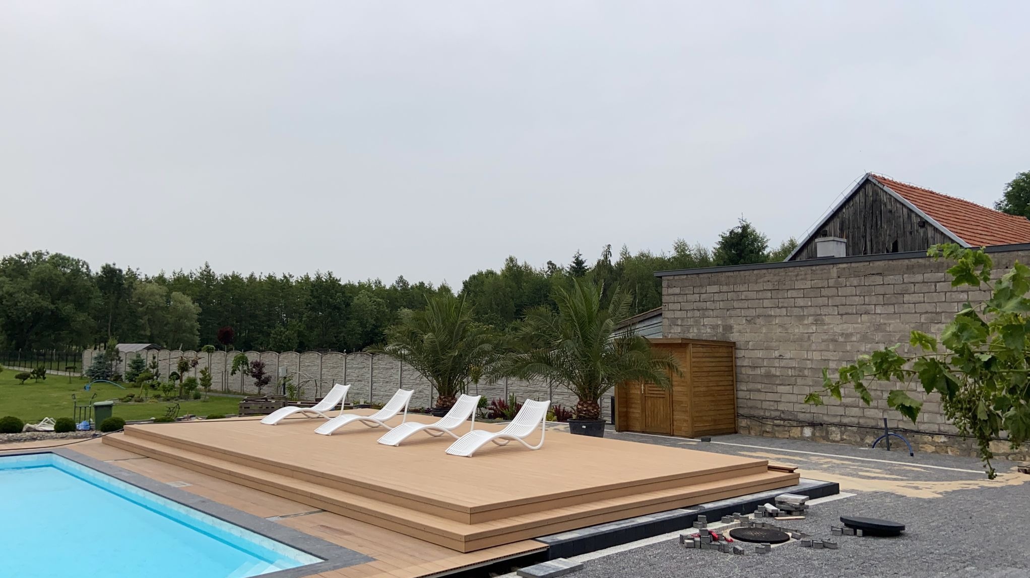 Nowoczesny basen z drewnianym tarasem i białymi leżakami, otoczony zielenią. W tle budynek z cegły i drzewa. Częściowo ukończone prace brukarskie.