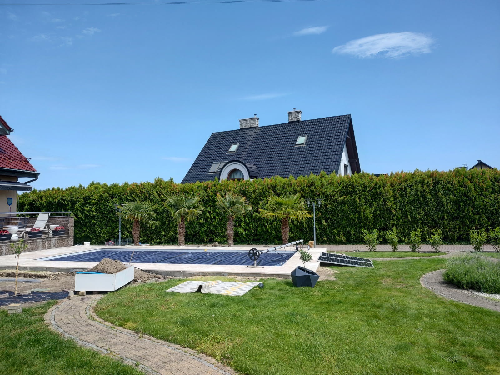 Zabezpieczony folią basen w ogrodzie otoczonym żywopłotem, z widocznymi palmami i domem w tle, w słoneczny dzień. Elementy przygotowania do sezonu.