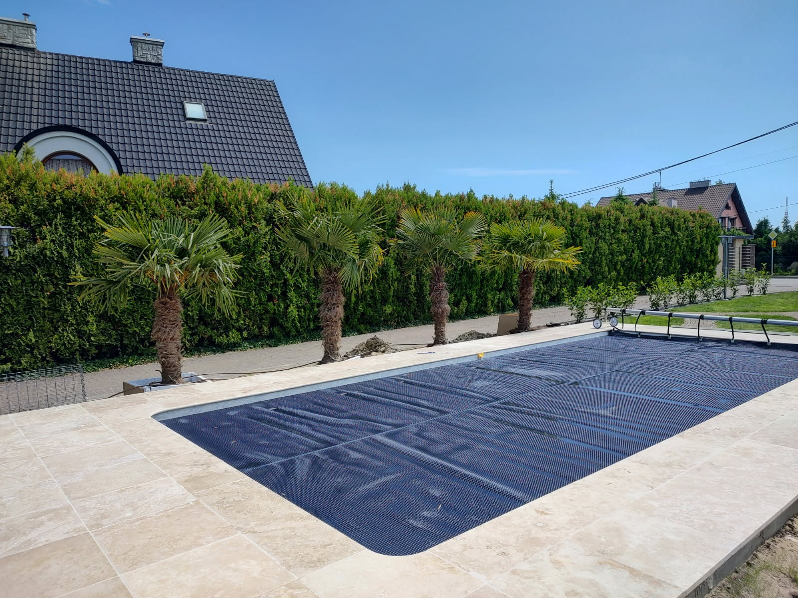 Zewnętrzny basen przy domu z pokrywą, otoczony palmami i zielonym żywopłotem pod błękitnym niebem. Wykończenie z jasnego kamienia.