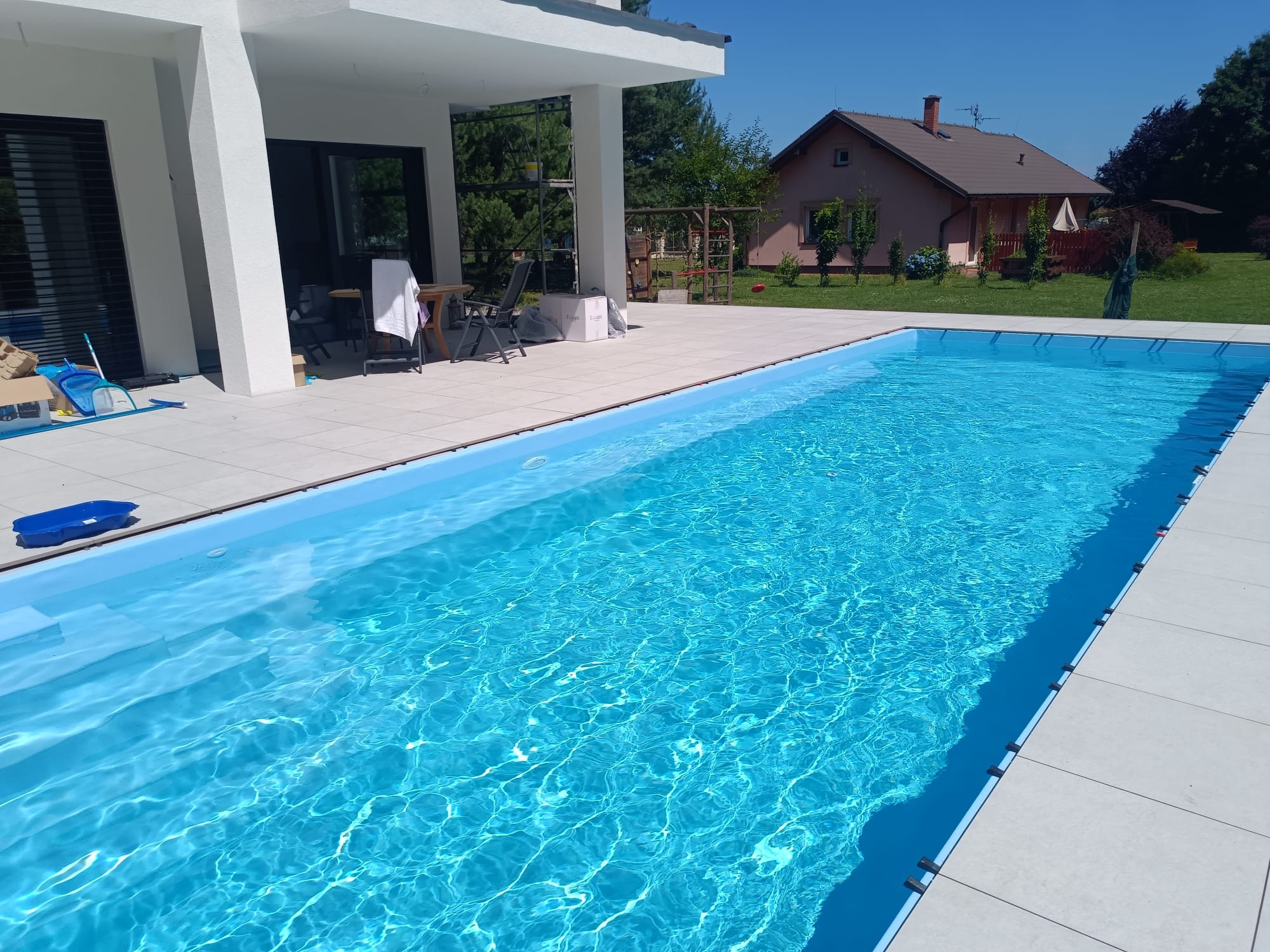 Błękitny, prostokątny basen z wodą w Tychach, otoczony jasnymi płytkami. W tle dom z ogrodem i błękitne niebo.