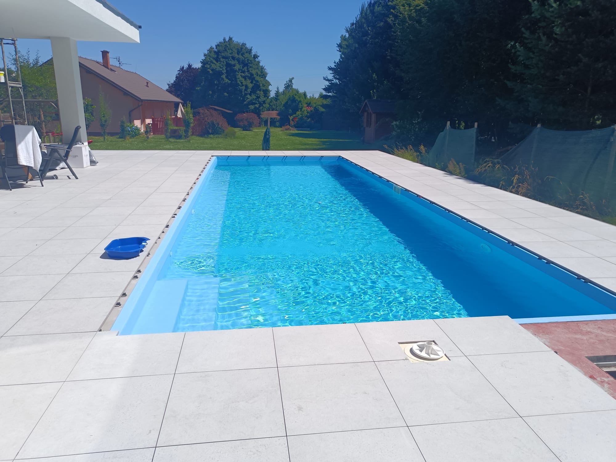 Błękitny basen z krystaliczną wodą otoczony jasnymi płytkami na zewnątrz domu w słoneczny dzień. Widoczne drzewa i budynek w tle.