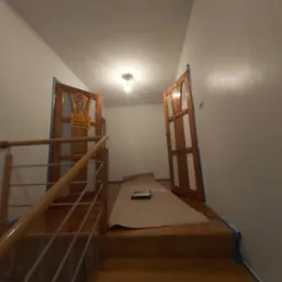 Przygotowanie do malowania ścian na klatce schodowej, zabezpieczone drewniane schody i ościeżnice drzwiowe folią malarską i taśmą, widoczny wałek do malowania.