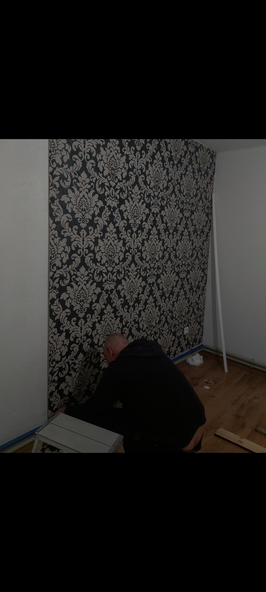 Mężczyzna w czarnej bluzie przykłada tapetę w ornamenty do ściany, stojąc na aluminiowym podnóżku w trakcie prac wykończeniowych.