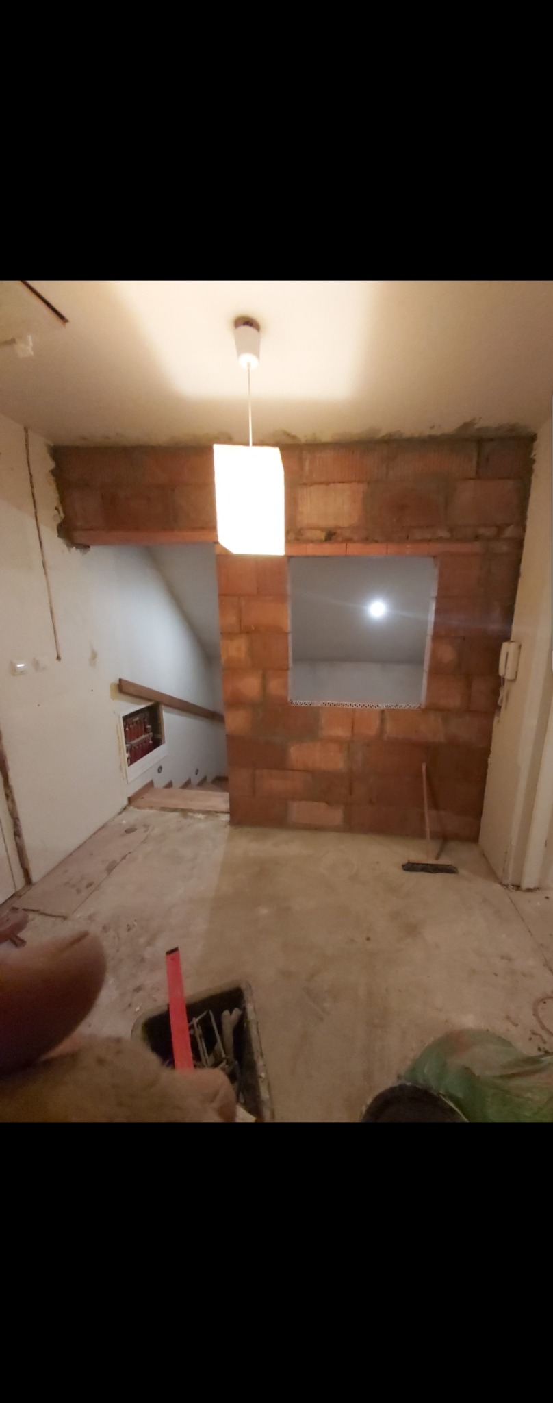 Wnętrze w trakcie budowy ścianki działowej z czerwonej cegły z otworem okiennym, widoczny surowy betonowy podkład, narzędzia w wiadrze na pierwszym planie i wisząca lampa.