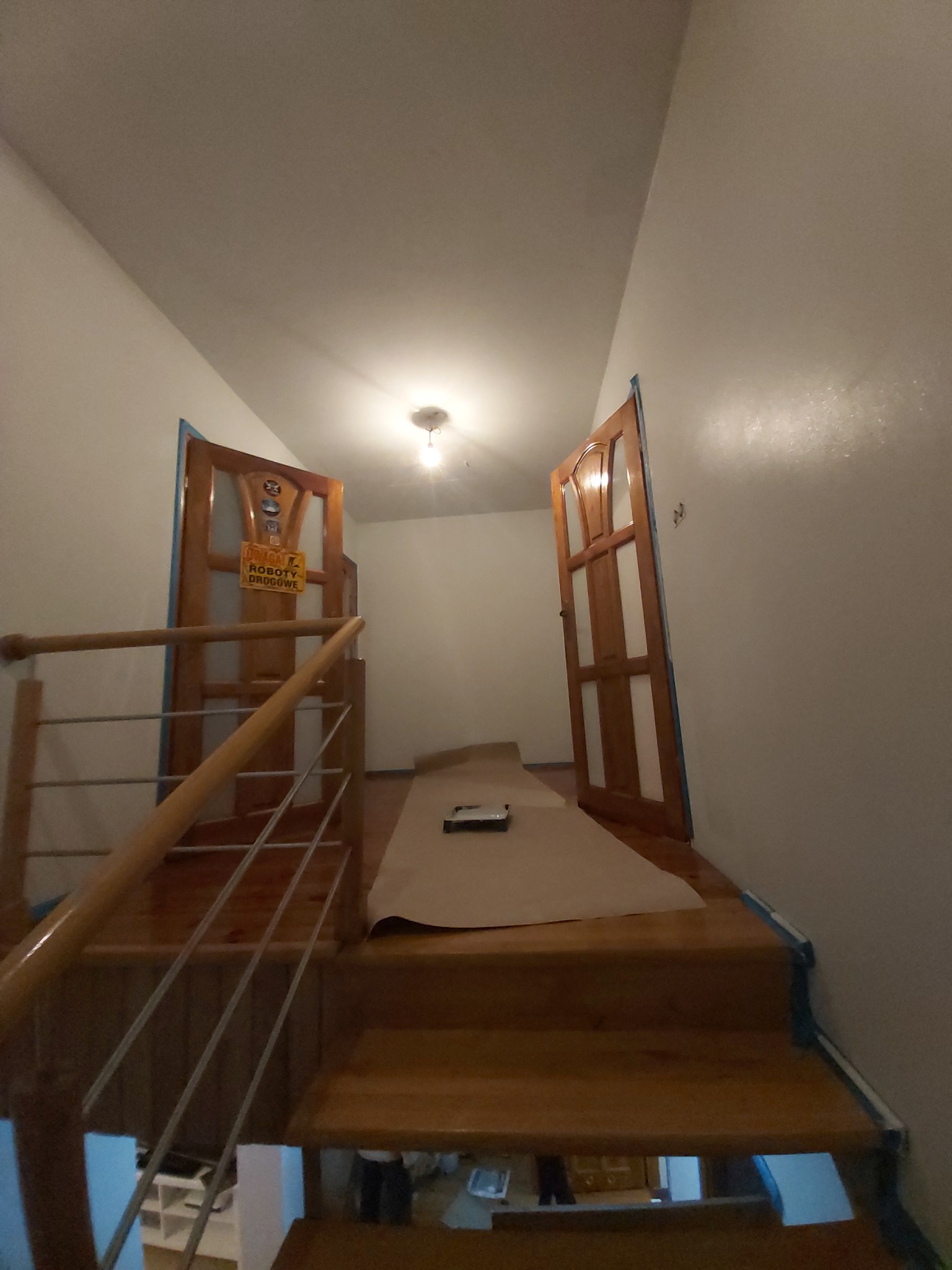 Przygotowanie do malowania ścian na klatce schodowej, zabezpieczone drewniane schody i ościeżnice drzwiowe folią malarską i taśmą, widoczny wałek do malowania.