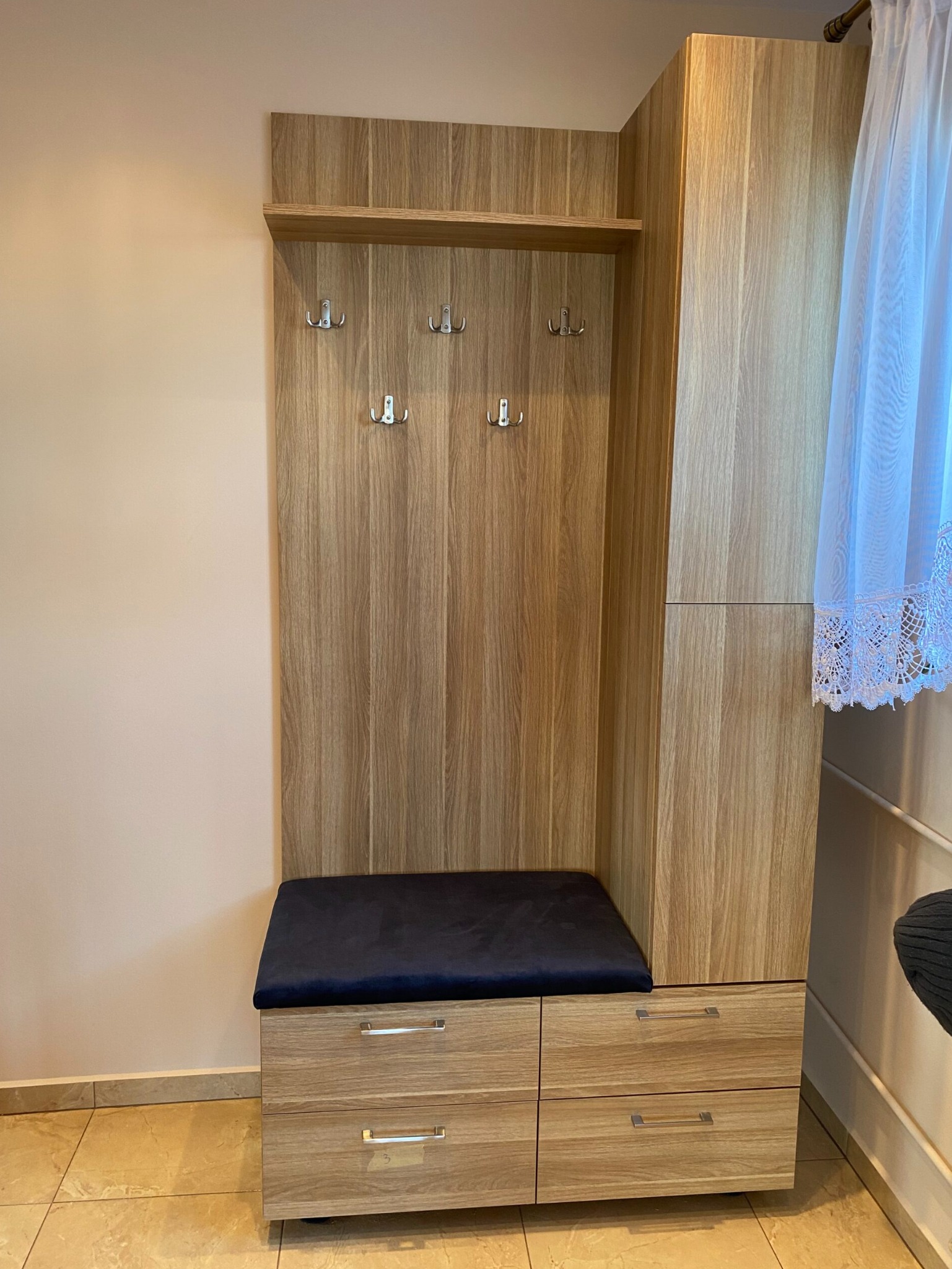 Drewniana zabudowa przedpokoju z wieszakami, półką i siedziskiem z granatową tapicerką, uzupełniona szafką z szufladami, na tle beżowej ściany i jasnej podłogi.