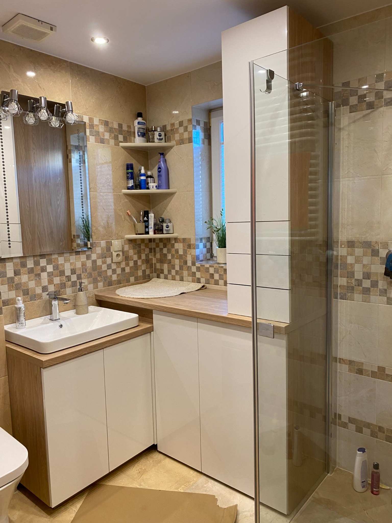 Nowoczesna łazienka z zabudową meblową pod umywalką, drewnianym blatem, kabiną prysznicową i płytkami mozaikowymi w odcieniach beżu i brązu.