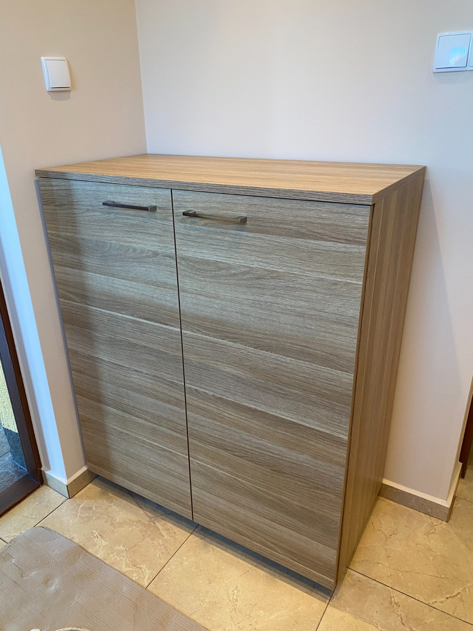 Dwudrzwiowa szafka z okleiną imitującą jasne drewno, z poziomym usłojeniem i metalowymi uchwytami, umieszczona przy ścianie w przedpokoju z beżowymi płytkami podłogowymi.