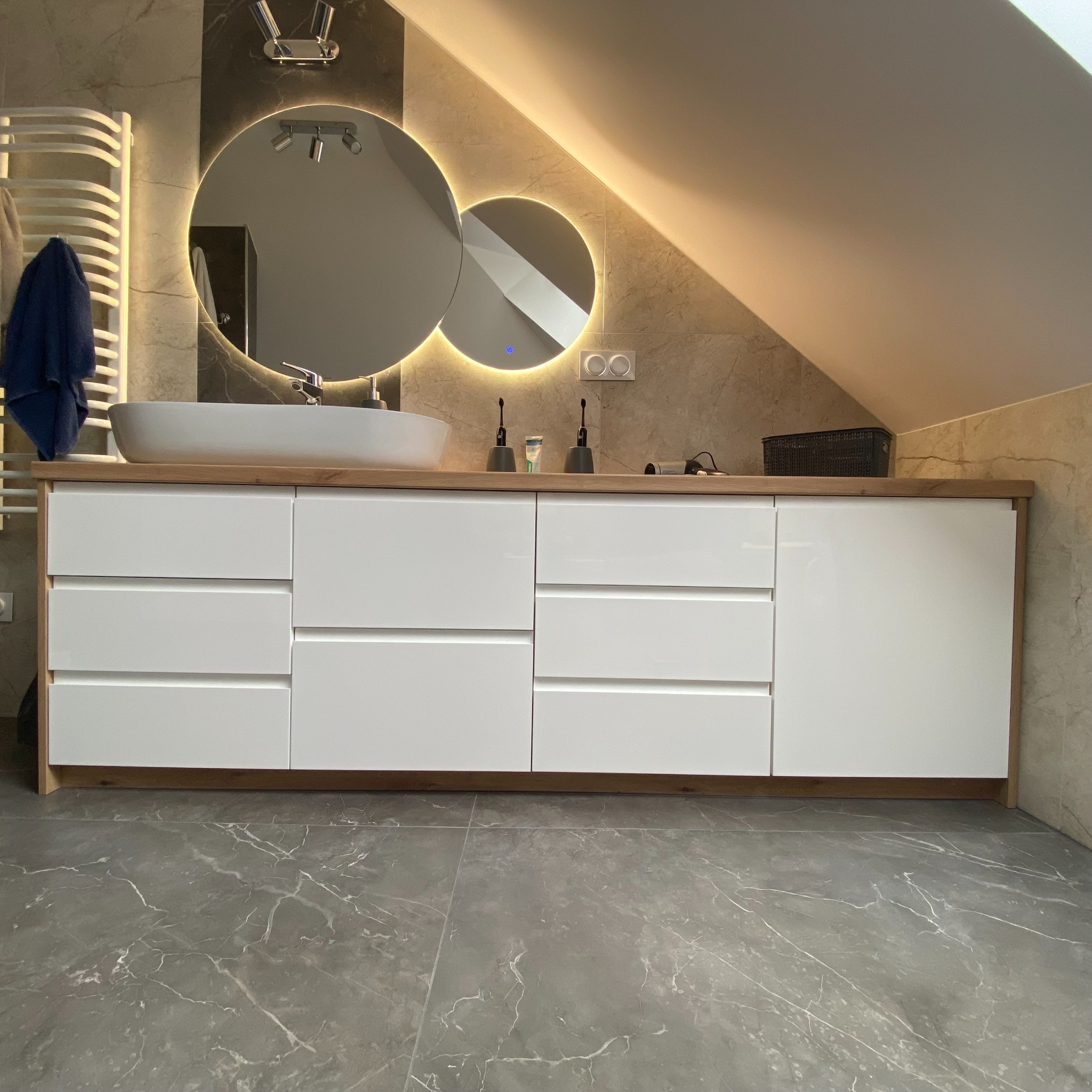 Nowoczesna łazienka na poddaszu z podświetlanym lustrem, białą umywalką nablatową i szafką z białymi frontami oraz drewnianym blatem i korpusem.