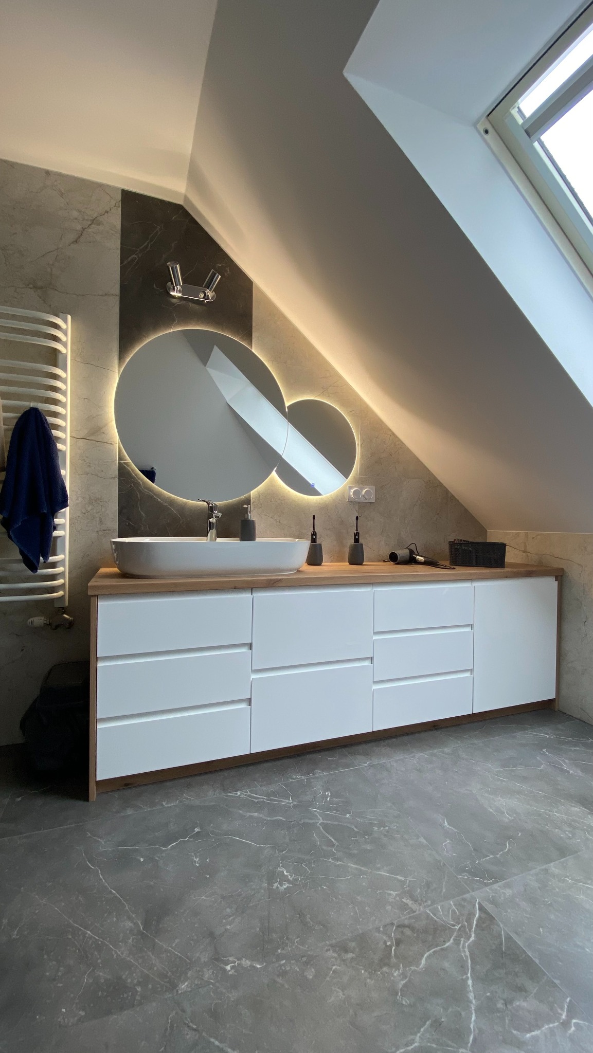 Nowoczesna łazienka na poddaszu z białymi szafkami, drewnianym blatem, owalną umywalką i podświetlanym lustrem w kształcie dwóch połączonych kół.