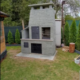 Murowany grill ogrodowy z szarej cegły na betonowej podstawie, z metalowymi drzwiczkami i rusztem, otoczony zielenią i drzewami.