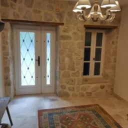 Kamienne ściany z oknem i przeszklonymi drzwiami. Wnętrze z dywanem, lampą i drewnianą ławką z kapeluszem. Widoczne elementy dekoracyjne.