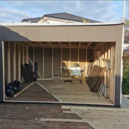 Wnętrze szkieletowej konstrukcji z drewnianym szkieletem, deskami podłogowymi i narzędziami. Widoczny fragment dachu domu w tle.