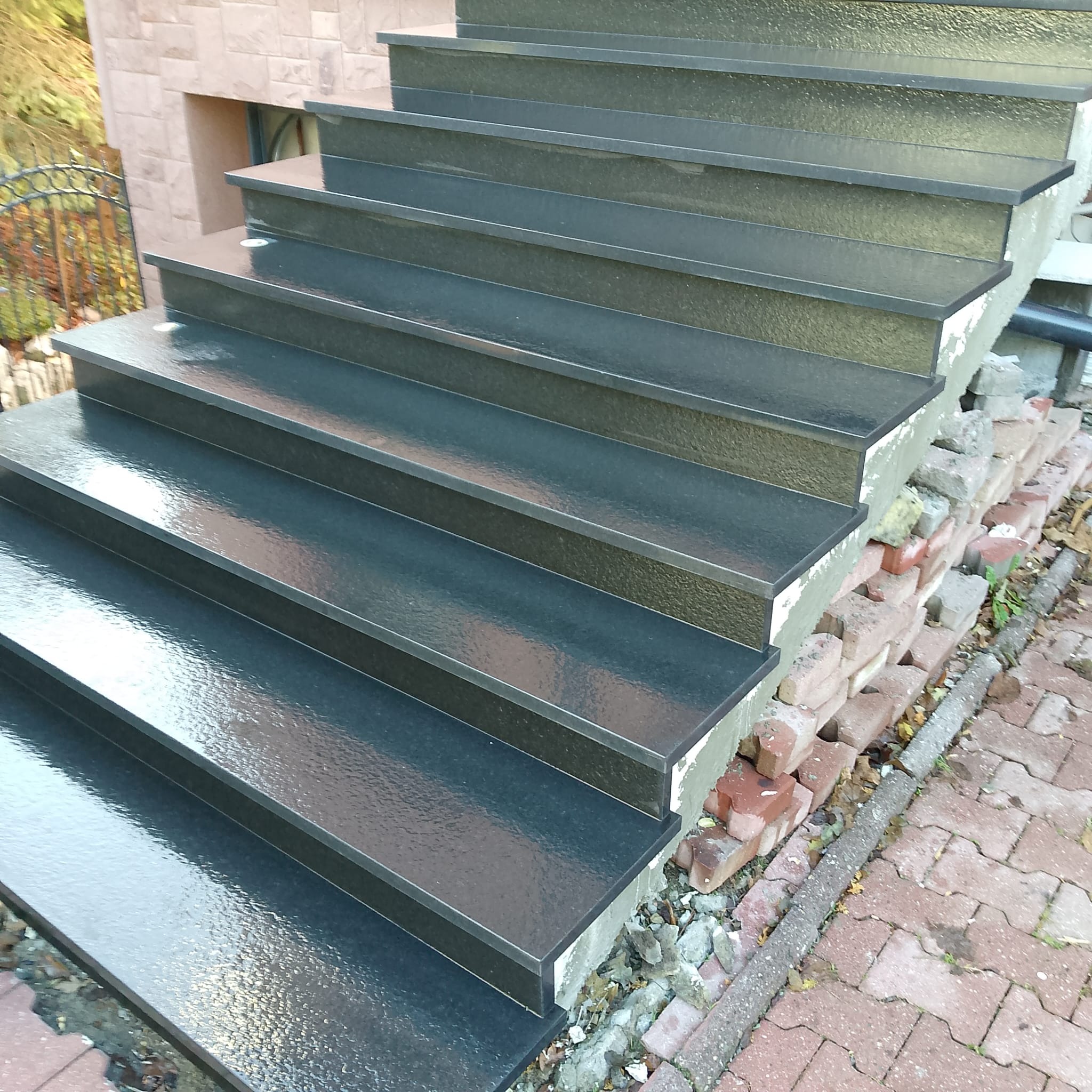 Zewnętrzne schody z ciemnego kamienia z połyskiem, prowadzące w górę. Widoczne detale wykończenia i otoczenie z kostki brukowej. Ujęcie z dołu.