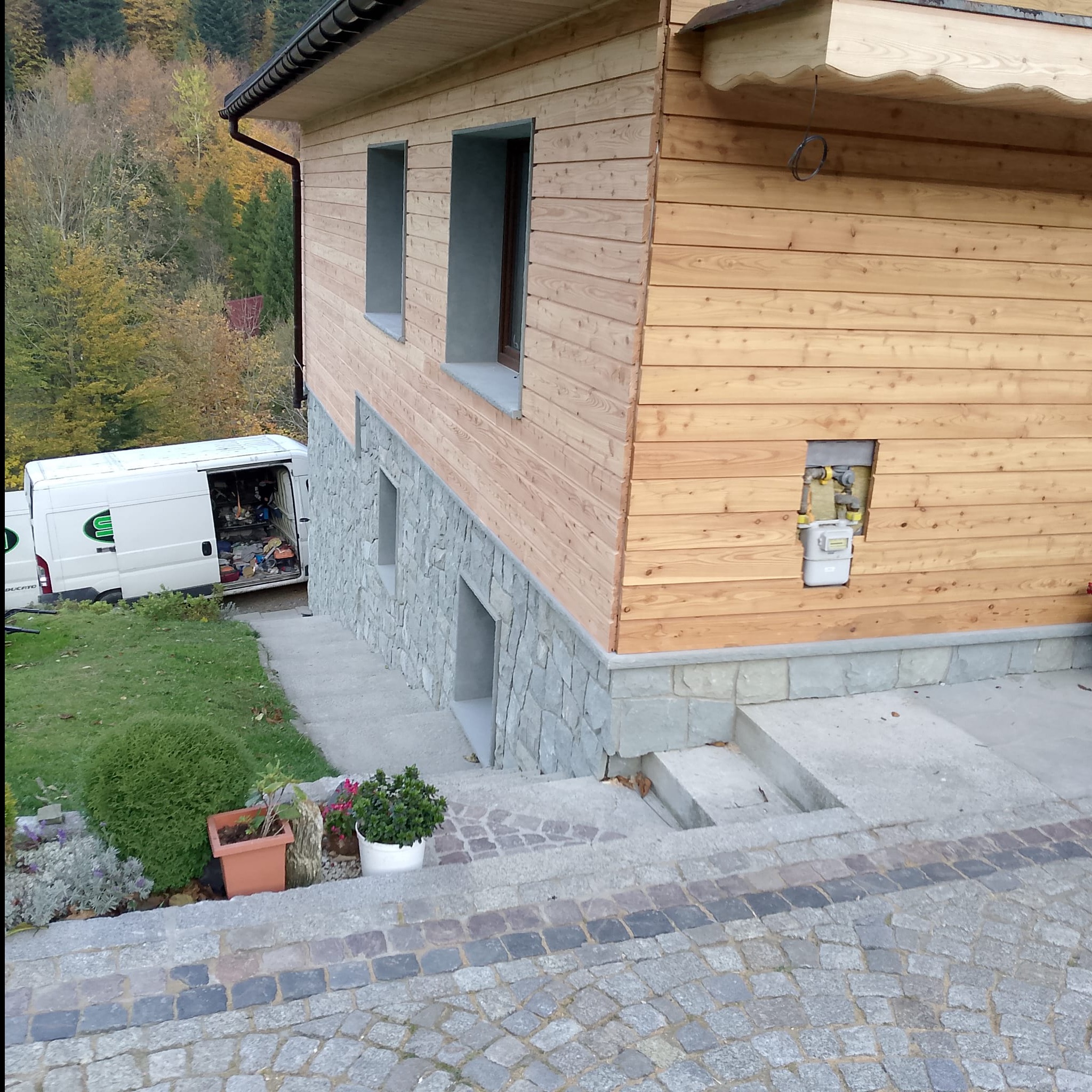 Dom szkieletowy z drewnianą elewacją i kamienną podmurówką. Widoczny fragment ogrodu i samochód dostawczy. Charakterystyczna zabudowa gazomierza.