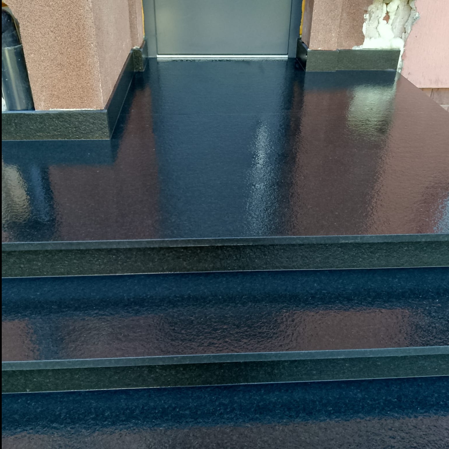 Czarne, błyszczące schody kamienne prowadzące do drzwi wejściowych, z widocznymi detalami wykończenia i odbiciami światła na powierzchni.