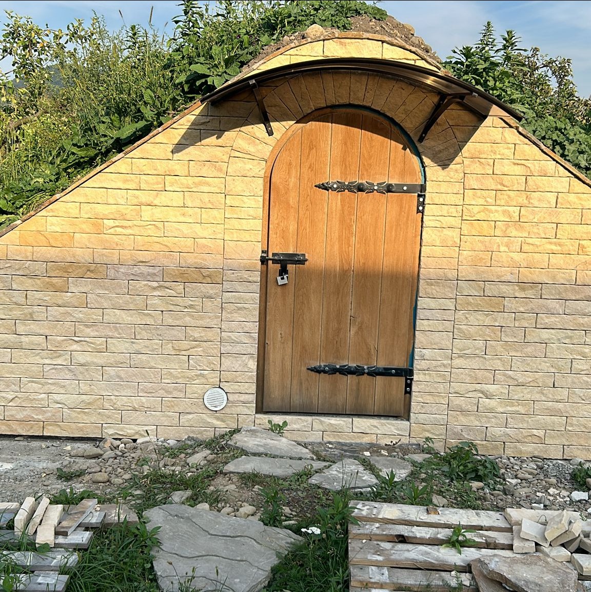 Zamurowane wejście do ziemianki z drewnianymi drzwiami i łukowym zadaszeniem, otoczone zielenią. Detale: ozdobne okucia, kamienna ścieżka.